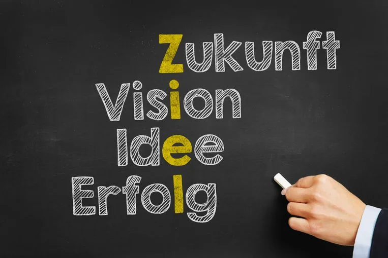 Hand schreibt auf eine Tafel die Wörter Zukunft, Vision, Idee und Erfolg, die Anfangsbuchstaben ergeben das Wort Ziel